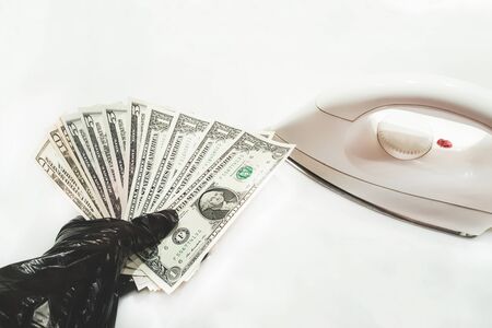 Hand in a black glove holds a bundle of money and gets ready to ironing cash - dollars bank notes -  with a hot iron for antibacterial disinfection. Clearing money. Isolated on white background. Panic and phobia  concepts caused by pandemic.の写真素材