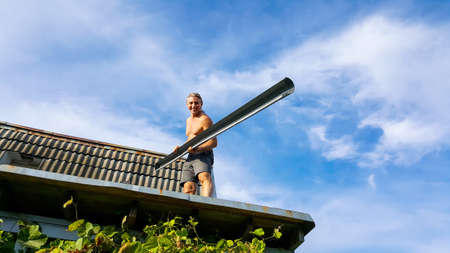 Down pipe installation by man-worker on the roof of the cottage. Isolated on the blue cloudy sky. Home and garden repair work in summer time.の写真素材