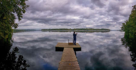 Man with long hair pulled back into a ponytail stands with his back on the pontoon of the river   Beautiful reflection of clouds on the water. Outdoor recreation and tourism concept. Adventure travel in Europe.の写真素材