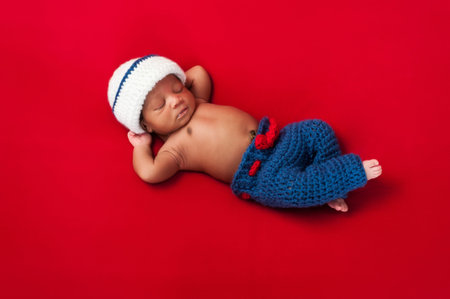 Five day old newborn baby boy wearing a red, white and blue sailor costume and sleeping on his back with his arms behind his head  Shot in the studio on a red background の写真素材