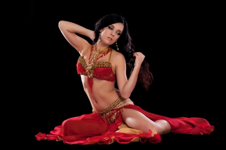 Full length portrait of a beautiful belly dancer wearing a red bellydance costume. She is sitting and posing with one hand behind her head. Isolated on black. の写真素材