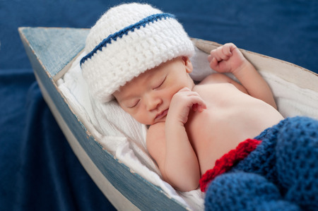 One week old newborn baby boy wearing a white and blue sailor hat. He is sleeping contentedly on his back in a little boat. の写真素材