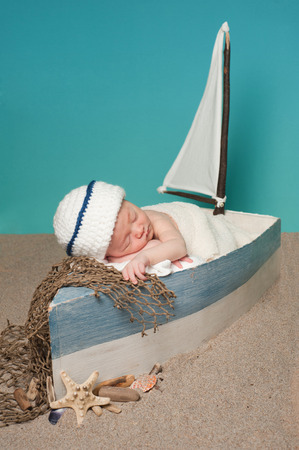 One week old newborn baby boy wearing a white and blue sailor hat. He is sleeping in a little sailboat surrounded by sand and seashells.の写真素材