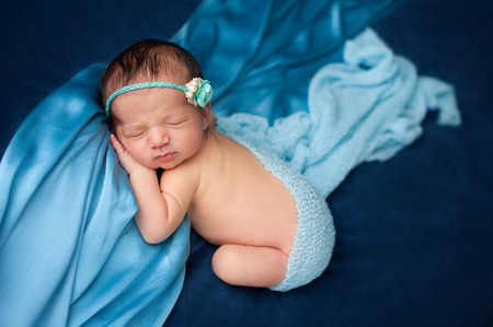 Six day old newborn baby girl wearing a paper rose and pearl headband and sleeping on blue fabric.の写真素材