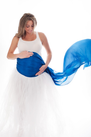 Beautiful pregnant woman wearing a white, full length tutu with a flowing, blue veil draped around her belly. Isolated on a white background.の写真素材