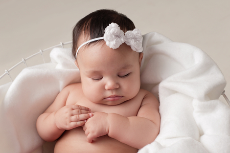 Four month old baby girl wearing a white, bow headband. She is sleeping on white fabric placed inside of a wire basket. Shot in the studio on a beige background.の写真素材