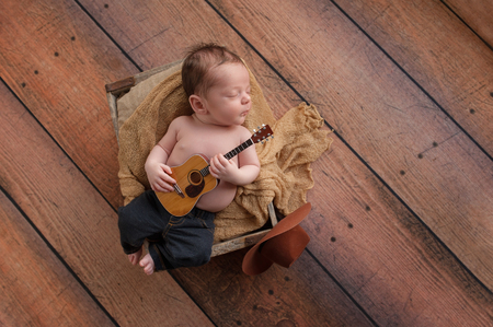 A three week old baby boy wearing jeans and playing a tiny acoustic guitar. He is lying in a wooden crate lined with burlap. Shot in the studio on a rustic, wood background.の写真素材