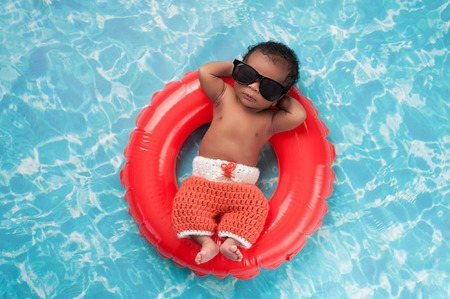 Two week old newborn baby boy sleeping on a tiny inflatable swim ring. He is wearing crocheted board shorts and black sunglasses.の写真素材