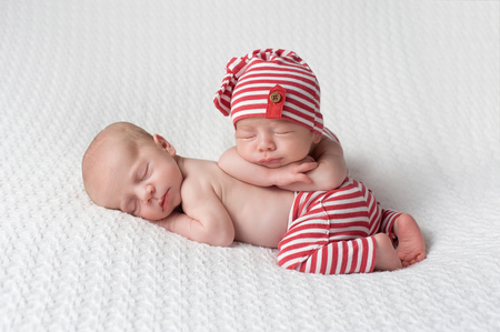 One month old fraternal, twin, newborn baby boys wearing red and white striped, up-cycled beanie and pants. They are stacked one on top of the other and sleeping on a white blanket.の写真素材