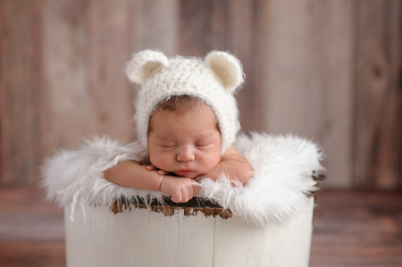 An 11 day old newborn baby girl sleeping in a little, wooden bucket. She is wearing a crocheted, white bear bonnet. Shot in the studio on a wood background.の写真素材