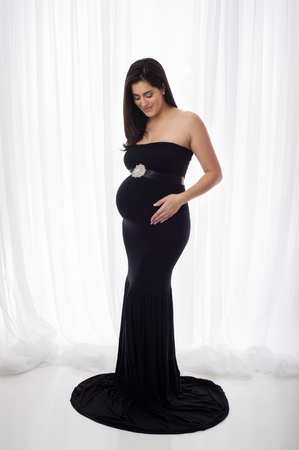Portrait of a beautiful pregnant woman wearing a black, fitted, mermaid style, full length gown. She is smiling and looking down at her belly.の写真素材