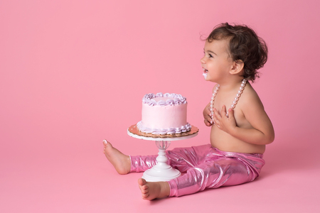 A happy, one year old, baby girl sitting with a cake. She is wearing pink leggings, a string of pearls and has frosting on her chin.の写真素材