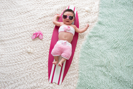 One month old baby girl lying on a tiny, pink and white surfboard. She is wearing sunglasses and a pink bathing suit.の写真素材