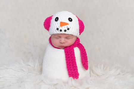 A newborn baby girl wearing a crocheted, bright pink and white snowgirl cap with matching neck scarf. Photographed on a white, flokati rug.の写真素材