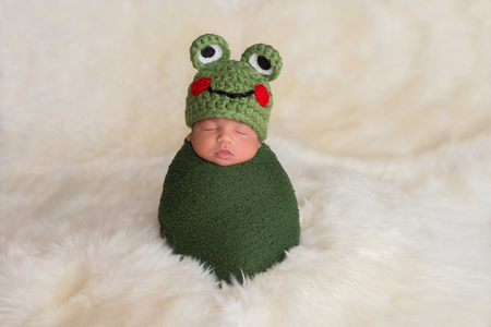Nine day old newborn baby boy wearing a green frog hat. He is sleeping upright while swaddled in a stretch wrap.の写真素材