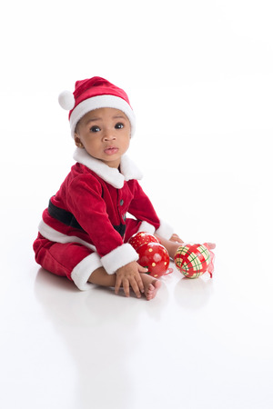 Six Month old baby girl wearing a Santa Claus costume. She is sitting on a white, seamless background with Christmas ornaments.の写真素材