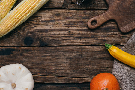 Autumn vegetables: pumpkins and corn with yellow leaves and cut board on a wooden background in kitchenの写真素材