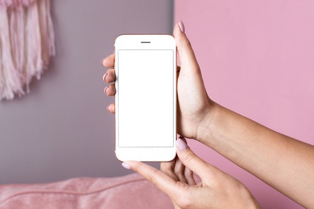 Female hands hold mobile phone with white screen mock up on a pink interior background side viewの写真素材