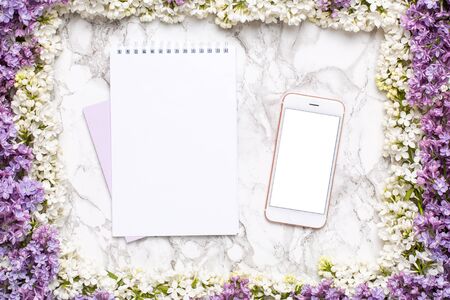 Mobile phone mock up, notebook and frame of white and lilac flowers on marble table in flat lay style top view.の写真素材