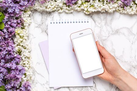 Mobile phone mock up in hand, notebook and frame of white and lilac flowers on marble table in flat lay style top view.の写真素材