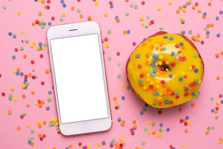 Mobile phone and sweet yellow donut on a pink background flat lay top viewの写真素材