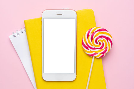 Mobile phone with a yellow notebook and colorful lolipop on a pink background top view