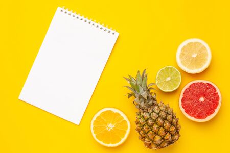 Notebook and tropical fruits pineapple, orange and lime on yellow background with copyspace top viewの写真素材