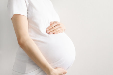 Pregnant woman in a white t-shirt holds her belly with her hands on a light background with...