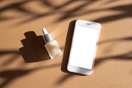 Liquid makeup foundation in glass bottle and mobile phone mock up on brown background with dark shadows , modern still life conceptの写真素材