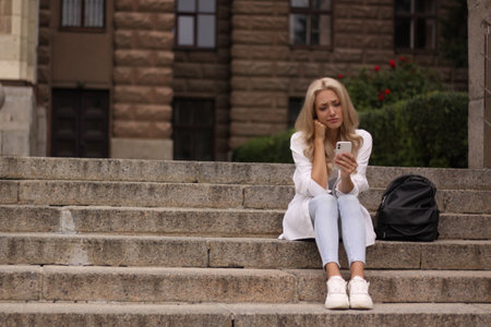 Young beautiful blonde girl in a white blouse and jeans sits on the steps in the city. She is upset about something.の写真素材