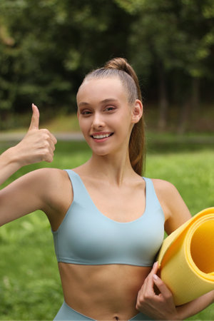 Sporty young woman holding yoga mat and showing thumbs up in parkの写真素材