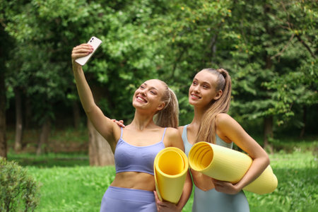 Two sporty women with yoga mats taking selfie on smartphone in parkの写真素材