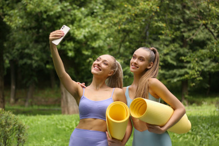 Two sporty women with yoga mats taking selfie on smartphone in parkの写真素材