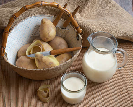 Vegan potato milk in a jug and glass and white raw potatoes in a wicker basket with a bamboo handle on a bamboo stand. natural background.の写真素材