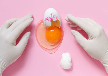 Hands in gloves and brocken egg with inscription EASTER over light pink background top view