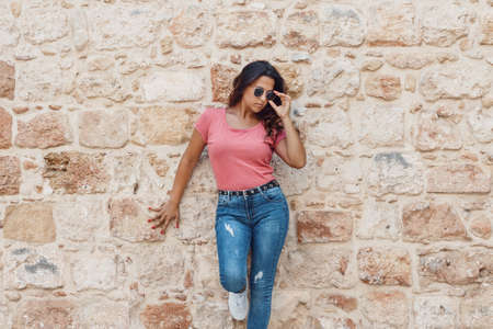Young women wearing t-shirt and jeans stays near a wall and touch sunglassesの写真素材