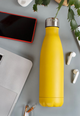 Yellow insulated bottle on grey desk surrounded by modern gadgets and plant in a vase  top viewの写真素材