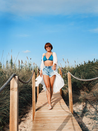 Young woman in cover up and jeans shorts walking between dunes.  Travel and vacations concept.