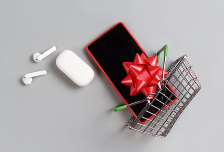 Smartphone, white wireless earphones and bow flying from the toy shopping cart top view on grey...