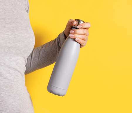 Close up of woman in grey tee holding grey insulated bottle on yellow backgroundの写真素材