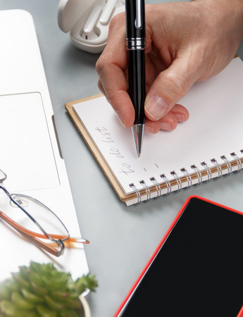 Man hand writing in note book on a grey office desk near laptop and cell phone close up