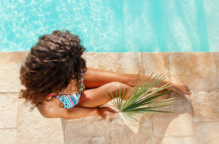Young caucasian woman in one-piece red swimsuit hides her face with tropical leaf beside a swimming pool top viewの写真素材