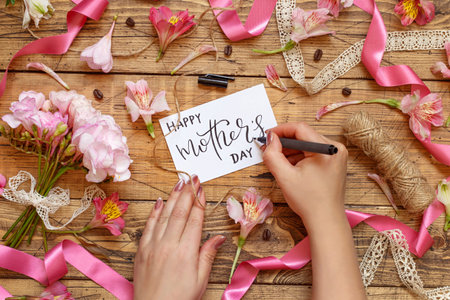 Hands Writing Happy Mother's Day card on a wooden table between pink flowers top view