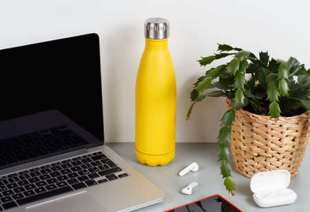 Yellow insulated bottle on grey desk surrounded by modern gadgets and plant in a vase close upの写真素材