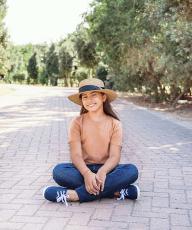 Smiling girl kid wearing t-shirt, jeans and straw hat sits outdoor in the parkの写真素材