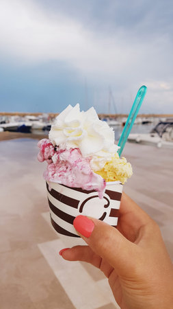 Woman's hand holding a cup with pink and yellow ice cream with whipped cream against the marina