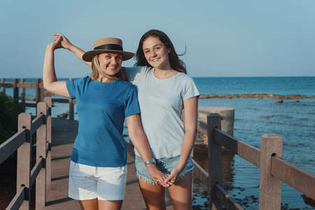 Mother and daughter standing on wooden walkway by the sea. A middle-aged woman in straw hat and teen girl holding each other hands, wearing blue t-shirts, smiling and looking straight into the cameraの写真素材