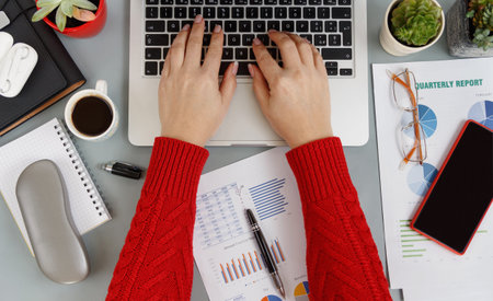 Woman hands wearing red sweater typing on laptop near reports, coffee and phone on grey office desk...