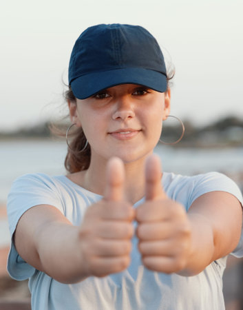 Teenage girl wearing blue baseball cap and showing big thumbs up near the sea at sunset. Head shot. Selective focus. Cap mockupの写真素材