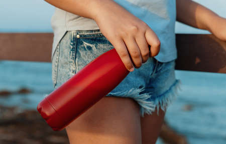 Young woman in light blue t-shirt holding red reusable water bottle in hand close up, cropped shot. Bottle and t-shirt mockupの写真素材